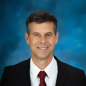 Professional headshot of a man with dark hair, wearing a black suit jacket, white shirt, and red tie, smiling against a blue background.