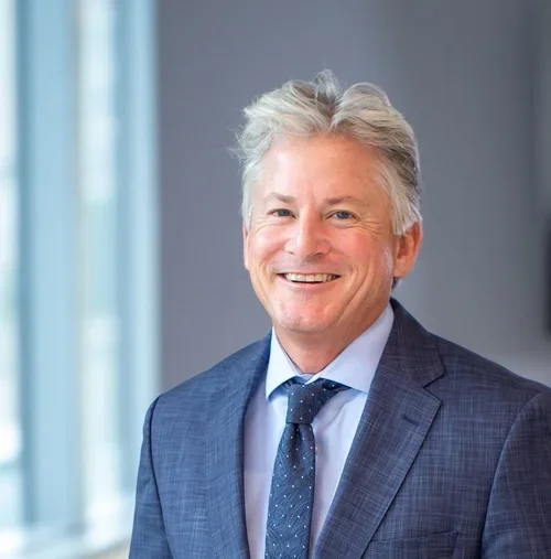 A smiling middle-aged man with gray hair, wearing a blue suit and tie, standing in a modern office with a window in the background.