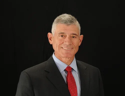 A middle-aged man with short gray hair, wearing a black suit, light blue dress shirt, and red tie, smiling against a black background.