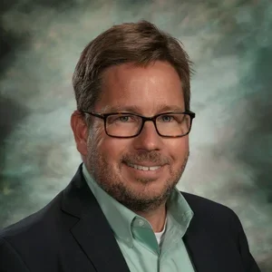 A professional headshot of a man with short brown hair, glasses, wearing a dark blazer and a light green shirt, smiling against a blurred background.