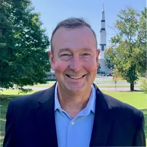 A smiling man in a suit standing outdoors with trees and a rocket in the background.