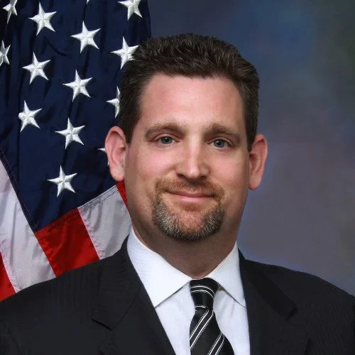 A man in a dark suit with a white shirt and a striped tie, standing in front of an American flag.