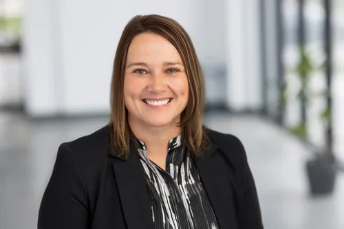 A smiling woman with shoulder-length brown hair wearing a black blazer and a black-and-white striped blouse, standing in a modern office space with large windows and potted plants in the background.