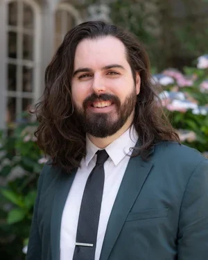A man with long dark hair and a beard smiling outdoors, wearing a dark suit jacket, white shirt, and black tie, with a background of greenery and a building with large windows.