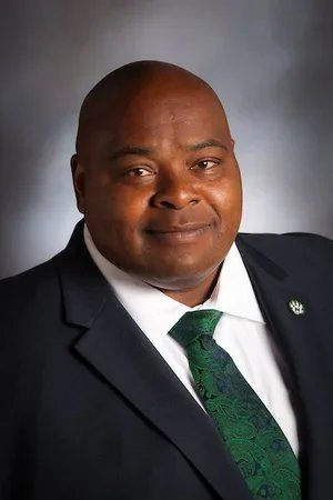 Professional headshot of a man wearing a dark suit, white shirt, and green patterned tie, against a gray background.