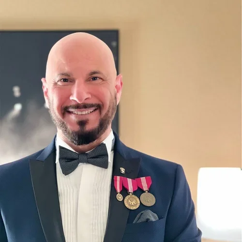 A smiling man with a bald head and a beard wearing a tuxedo and bow tie, with three medals pinned to his jacket, standing indoors.