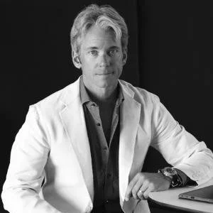 Black and white portrait of a middle-aged man with light hair, wearing a white blazer and a dark shirt, sitting at a desk with a laptop, against a dark background.
