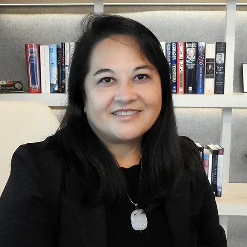 A woman with black hair and a necklace, sitting in front of a bookshelf filled with books.