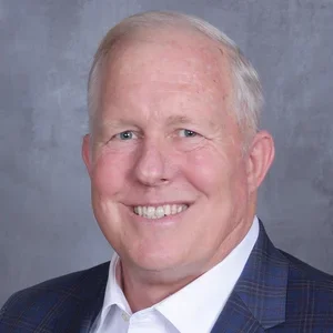 A smiling man with short white hair wearing a dark blazer over a white shirt, against a gray background.