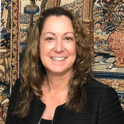 A woman with long, wavy brown hair smiling at the camera, wearing a black jacket, with a colorful tapestry featuring floral and patterned designs in the background.