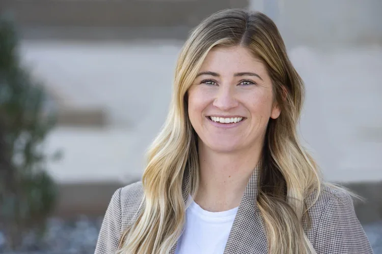 Headshot of a smiling woman with long wavy blonde hair, wearing a gray blazer and white shirt, outdoors in a bright setting.