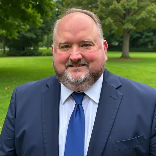 Middle-aged man in a navy suit and blue tie smiling outdoors with trees and grass in the background.