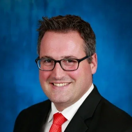 A man with short dark hair, wearing glasses, a black suit, white shirt, and red tie, smiling against a blue background.