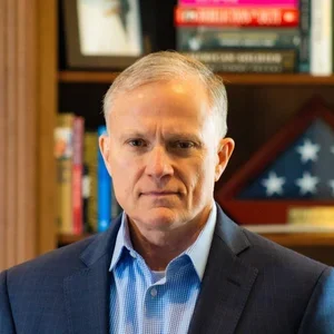 A middle-aged man with gray hair, wearing a dark blazer and light blue shirt, standing in front of a bookshelf with books and a folded American flag.