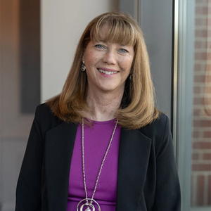 A woman with light brown hair and bangs, wearing a black blazer over a purple top, smiling in front of a window.
