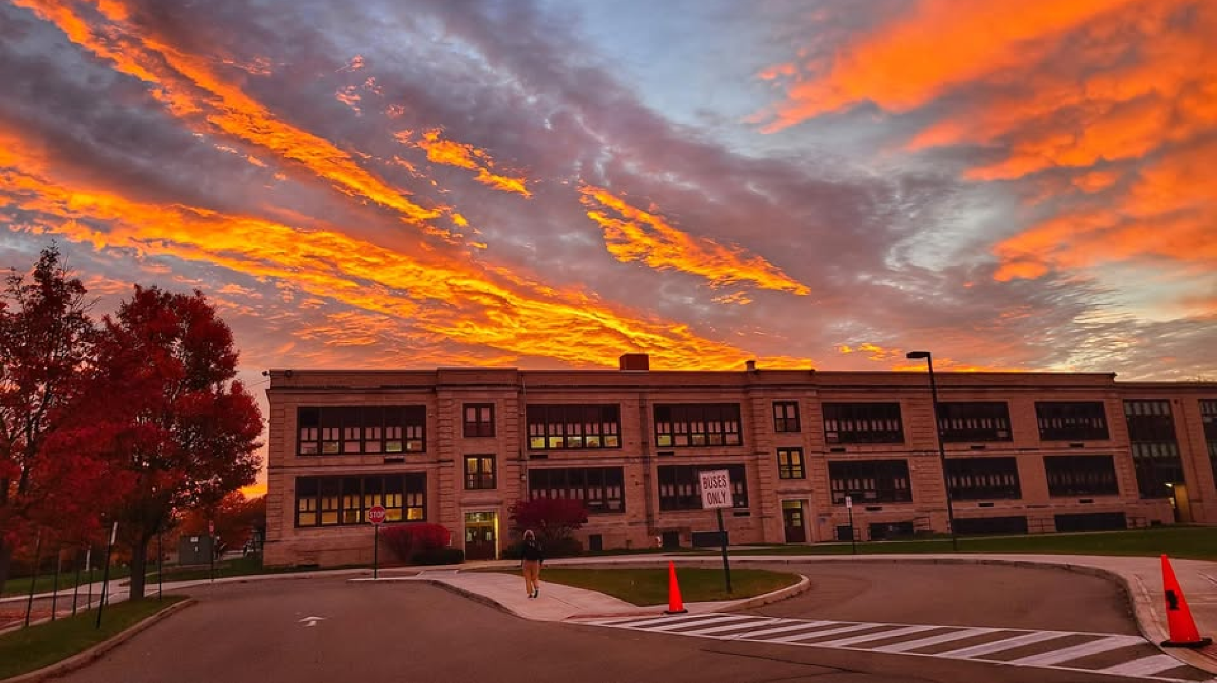 Sunset sky with vibrant orange and yellow clouds above a school building with trees on the left and a student walking on the sidewalk.