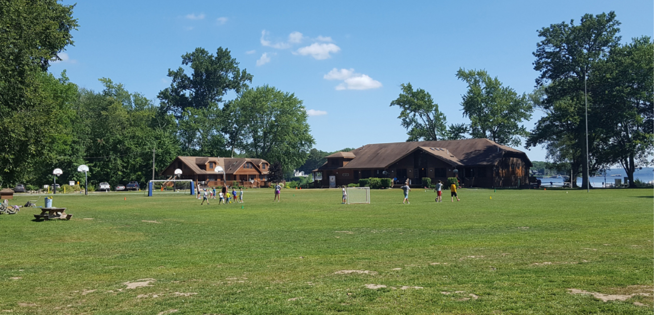Children playing soccer on a grassy field near buildings and trees, with a lake visible in the background on a sunny day.