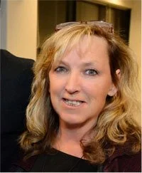 A woman with long wavy brown hair and glasses on her head, wearing a blue plaid shirt, smiling indoors with framed pictures and a black-and-white photograph behind her.