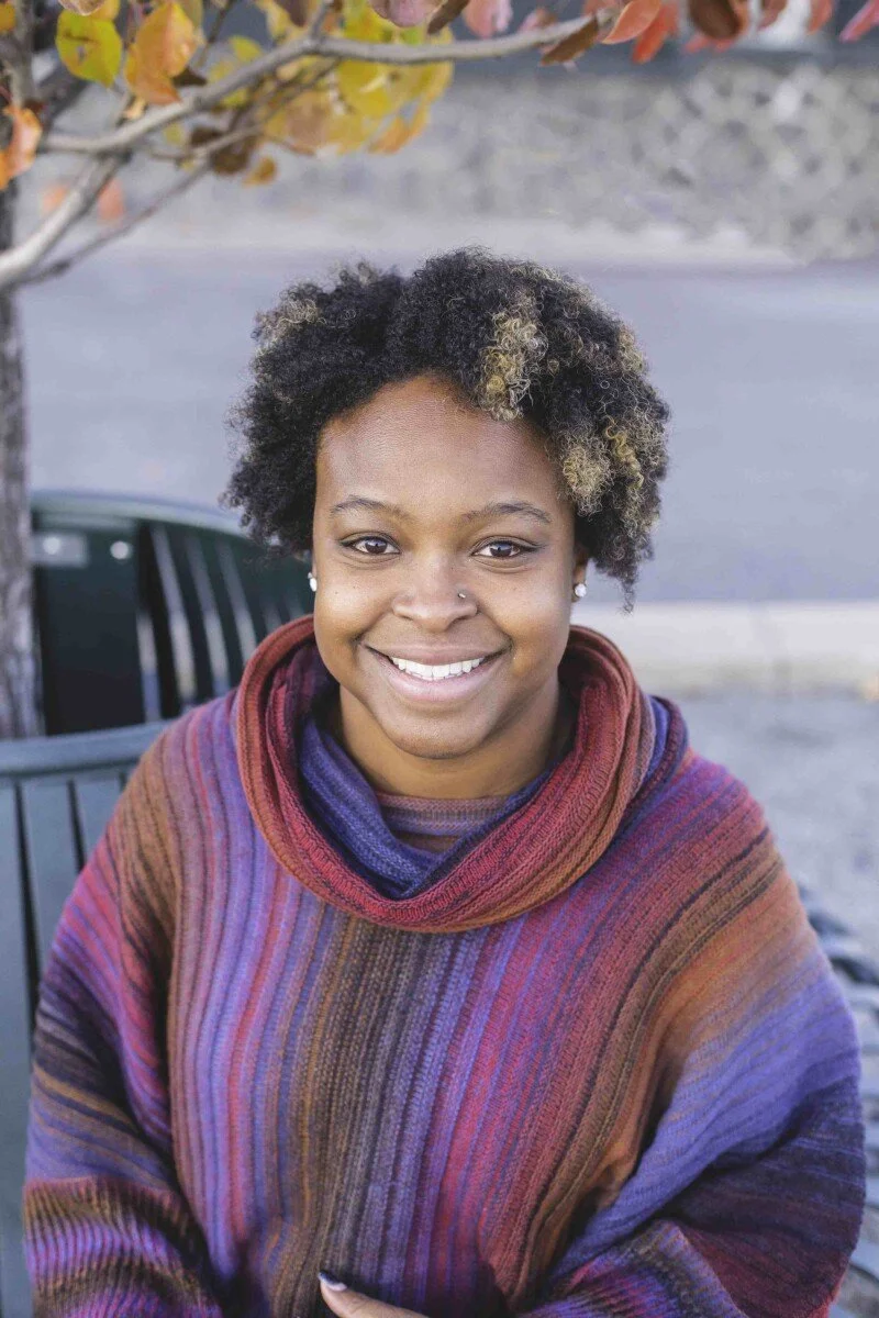 A smiling woman with curly hair sitting outdoors in front of a tree with autumn leaves, wearing a colorful striped sweater.