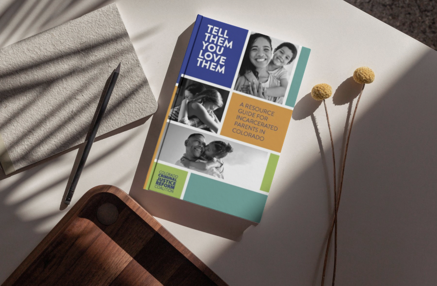 Book titled 'Tell Them You Love Them' with subtitle 'A Resource Guide for Incarecerated Parents in Colorado,' placed on a white surface with a gray notebook, a black pencil, and three yellow flowers in shadow.