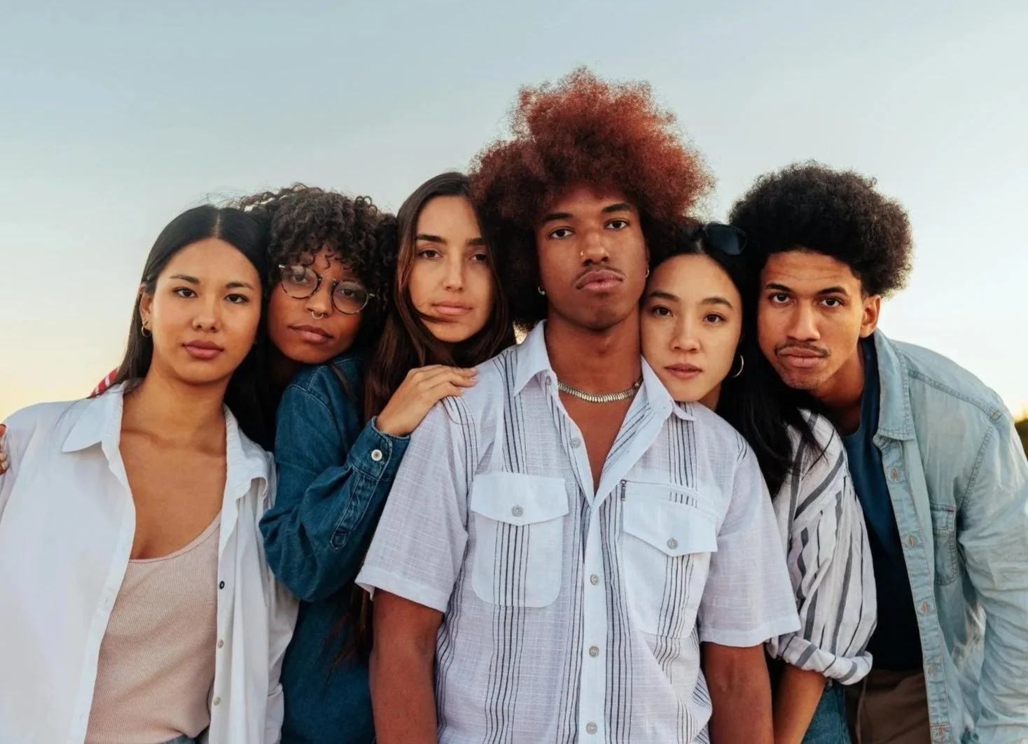 Group of six diverse young adults standing close together outdoors during sunset.