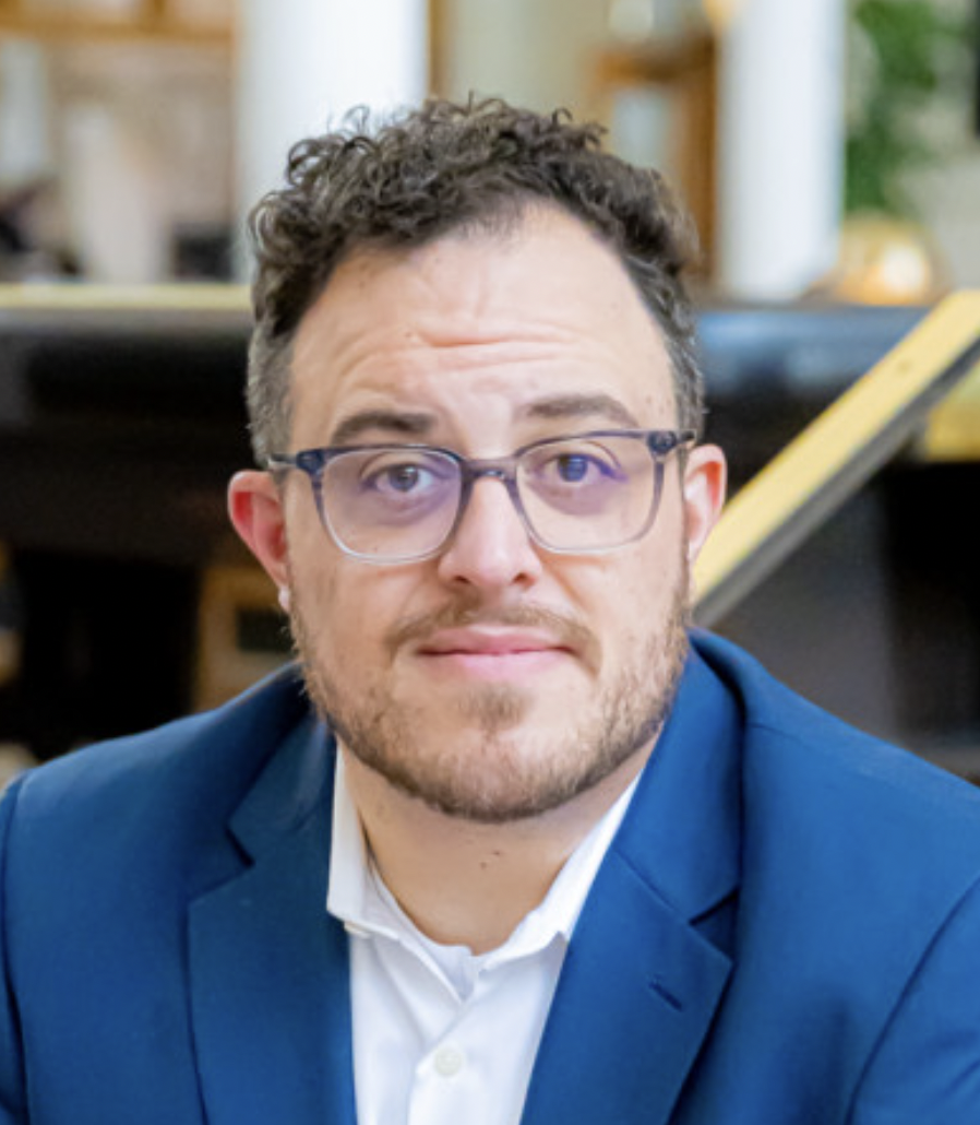 A man with glasses and a beard wearing a blue suit and white shirt, sitting indoors with a blurred background.