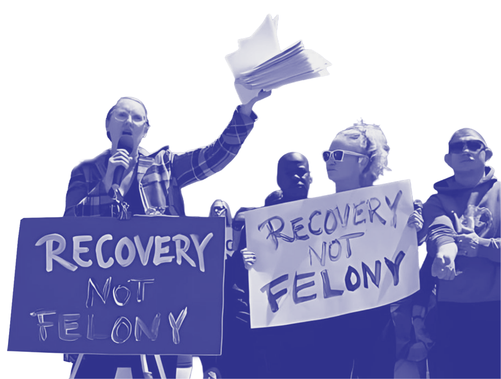 People protesting for drug policy reform holding signs that read 'Recovery Not Felony' and 'Recovery Not Felony', with one person speaking into a microphone and others clapping and holding signs.