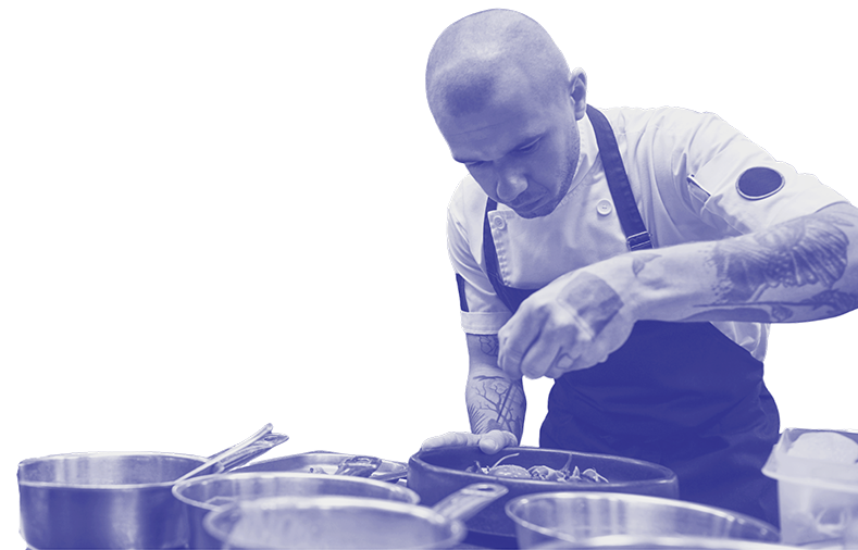 A chef with tattoos wearing a white uniform and apron, focused on preparing food in a kitchen.