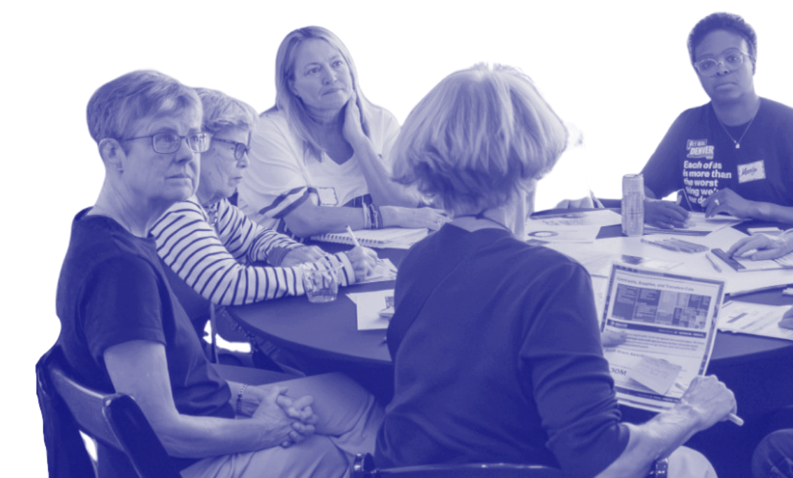 Group of women sitting around a table engaged in discussion, with papers and a tablet in front of them.