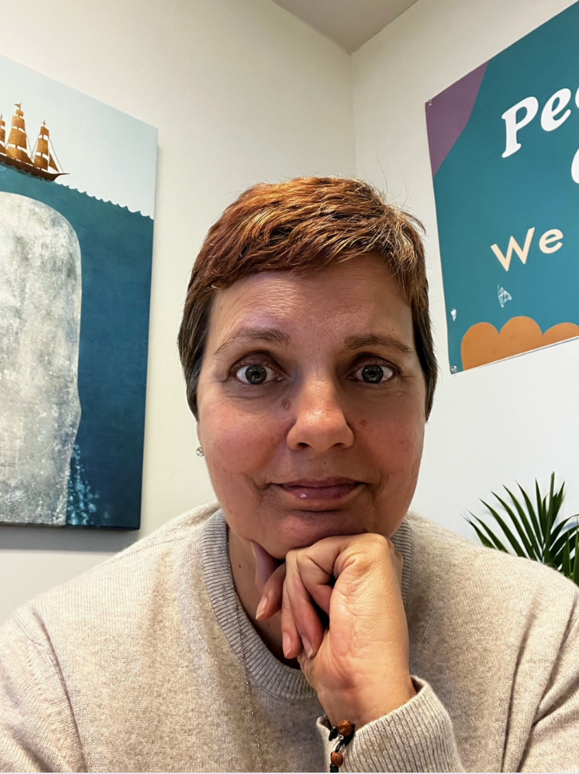 A woman with short brown hair sitting in an indoor space, resting her chin on her hand, wearing a beige sweater with a beaded bracelet, with artwork and a sign partially visible behind her.