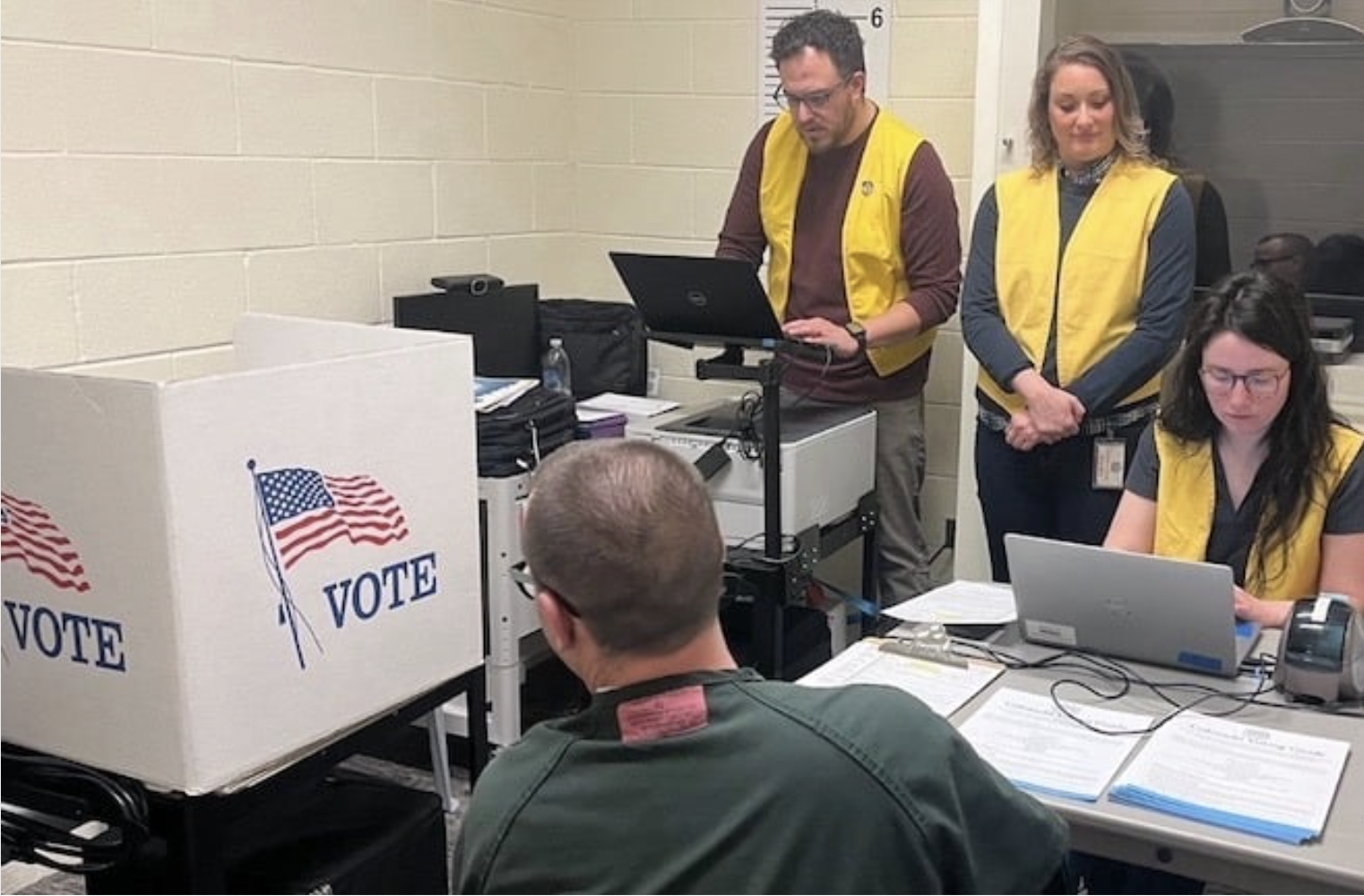 People working at a voting station with ballot boxes labeled 'VOTE', some using laptops and others standing or sitting, in a room with beige walls.