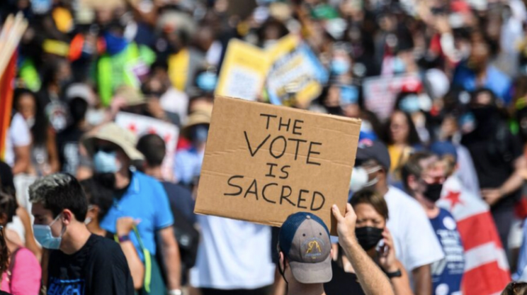A large crowd of people wearing masks at a demonstration, with one person holding a cardboard sign that reads, "The vote is sacred."