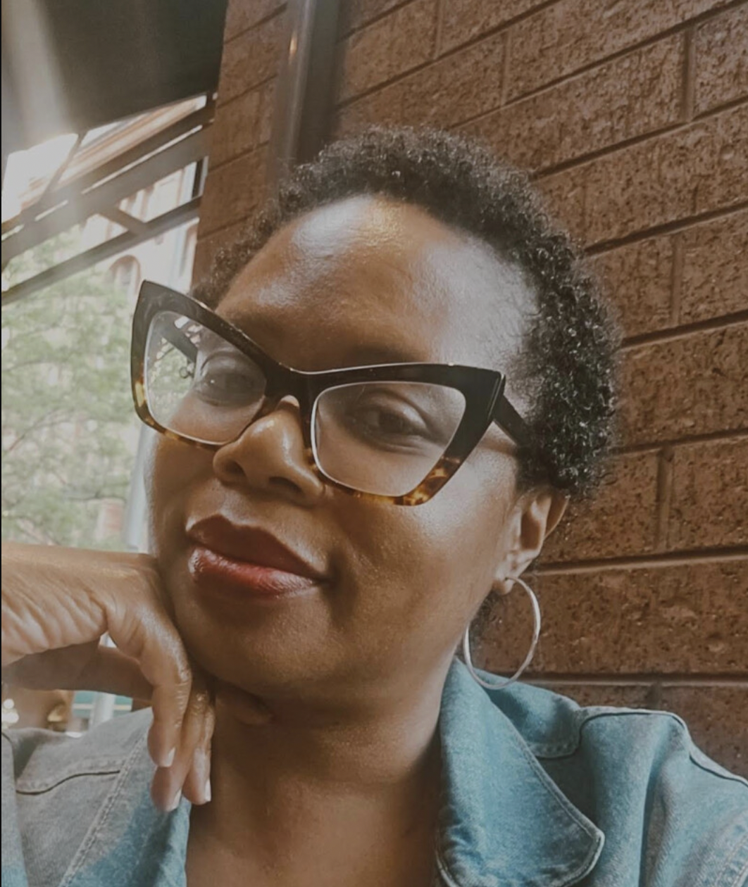 Close-up of a woman wearing glasses and a denim jacket, sitting against a brick wall inside a cafe or restaurant.