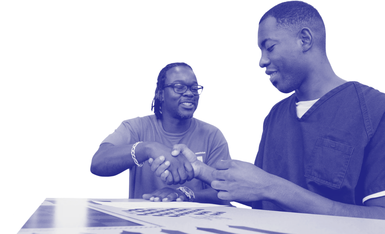 Two men shake hands over a table, one wearing glasses and the other wearing scrubs, in a friendly interaction.