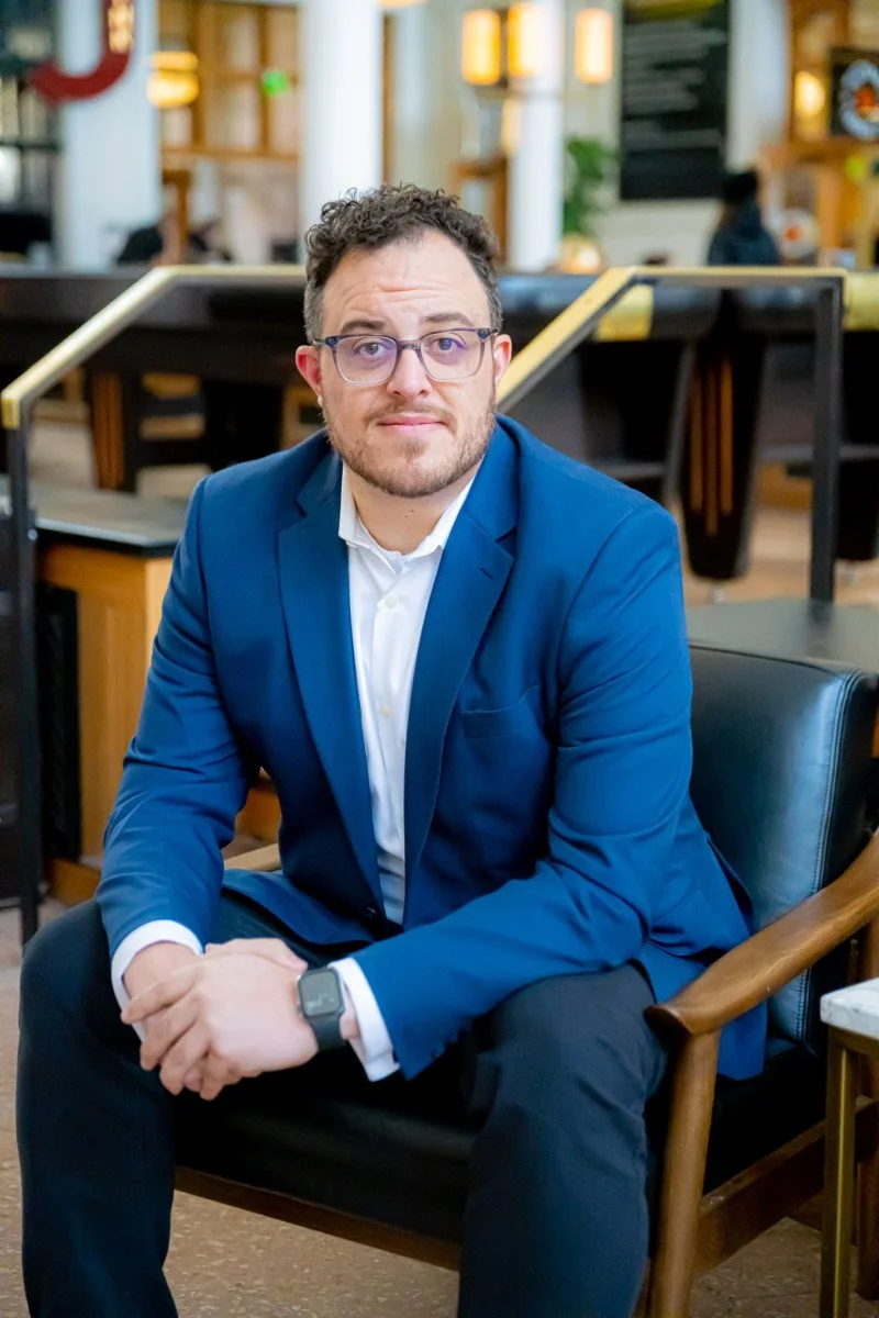 A man in a blue suit with glasses sitting on a chair in a modern, brightly lit indoor space, possibly a cafe or restaurant.