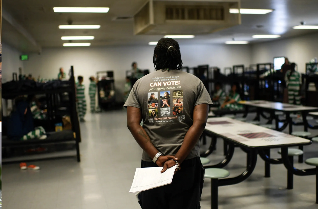 Person standing inside a large room, possibly a detention or quarantine center, with several children in striped clothing in the background. The individual is wearing a gray t-shirt with voting advocacy messages and holds a paper in their hand.
