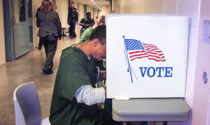 Person casting a ballot at a voting booth with an American flag and the word 'VOTE' on it, in an indoor polling station.