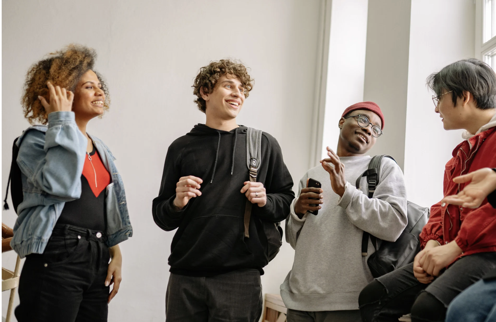 Group of four diverse young adults standing and talking indoors near large windows, smiling and engaging with each other.