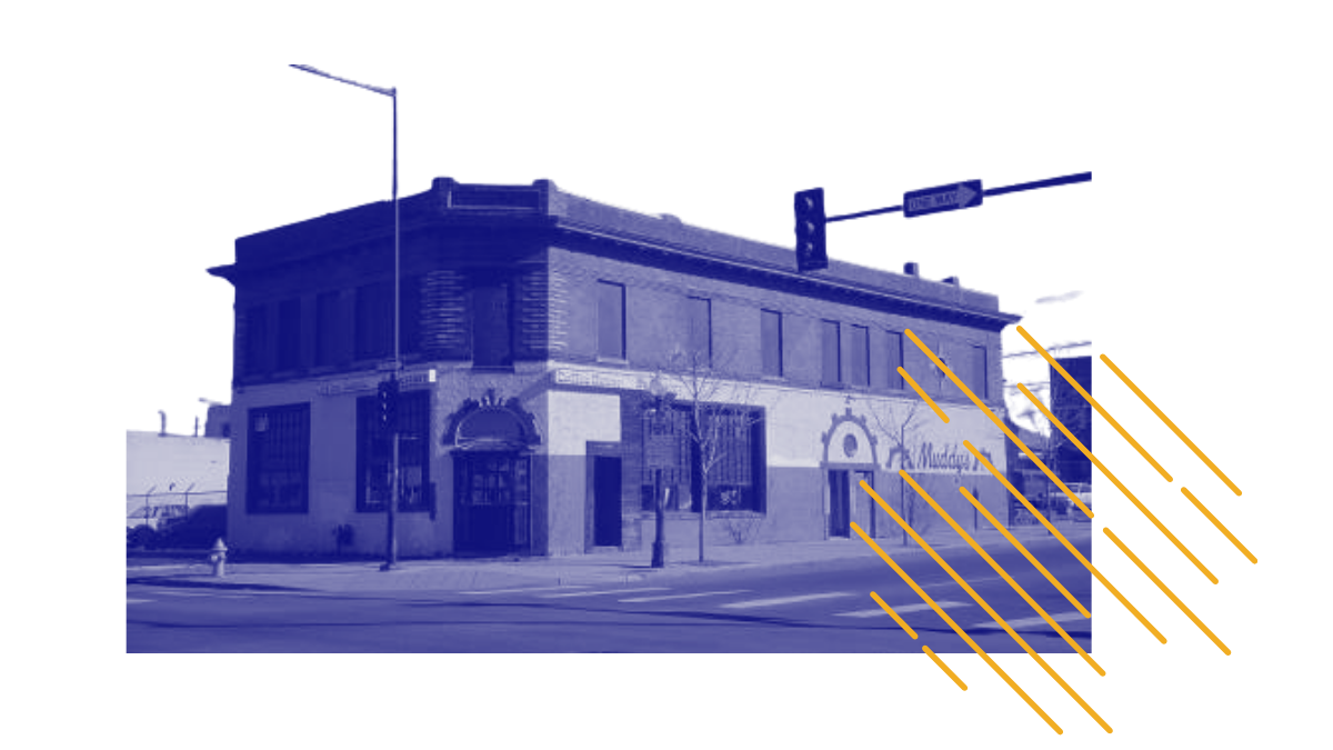 A corner building with large windows and a sign that reads 'The Muddah.' There's a fire hydrant and a street lamp in front, with a crosswalk and traffic signals on the corner.