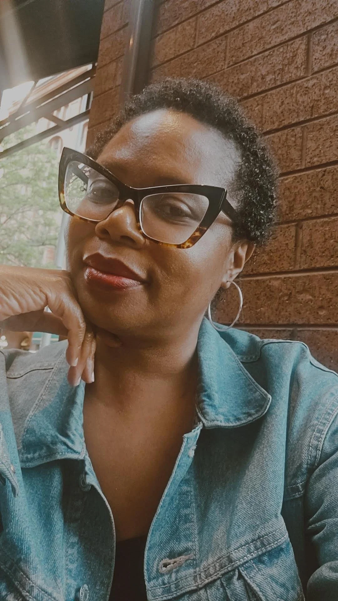 Close-up of a woman wearing large glasses and hoop earrings, smiling while sitting in front of a brick wall at a cafe.
