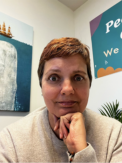 A woman with short brown hair sitting indoors, resting her chin on her hand, with colorful wall art and a plant in the background.
