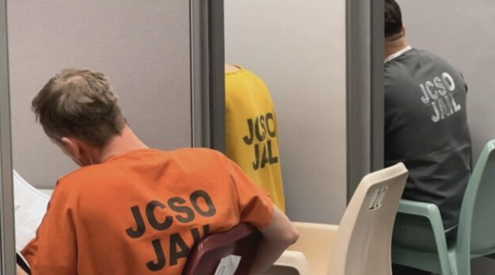 Two incarcerated people sitting with their backs turned in a jail visitation room. The orange jumpsuit has 'JCSO JAIL' printed on it, and the gray jumpsuit has the same text on its back.