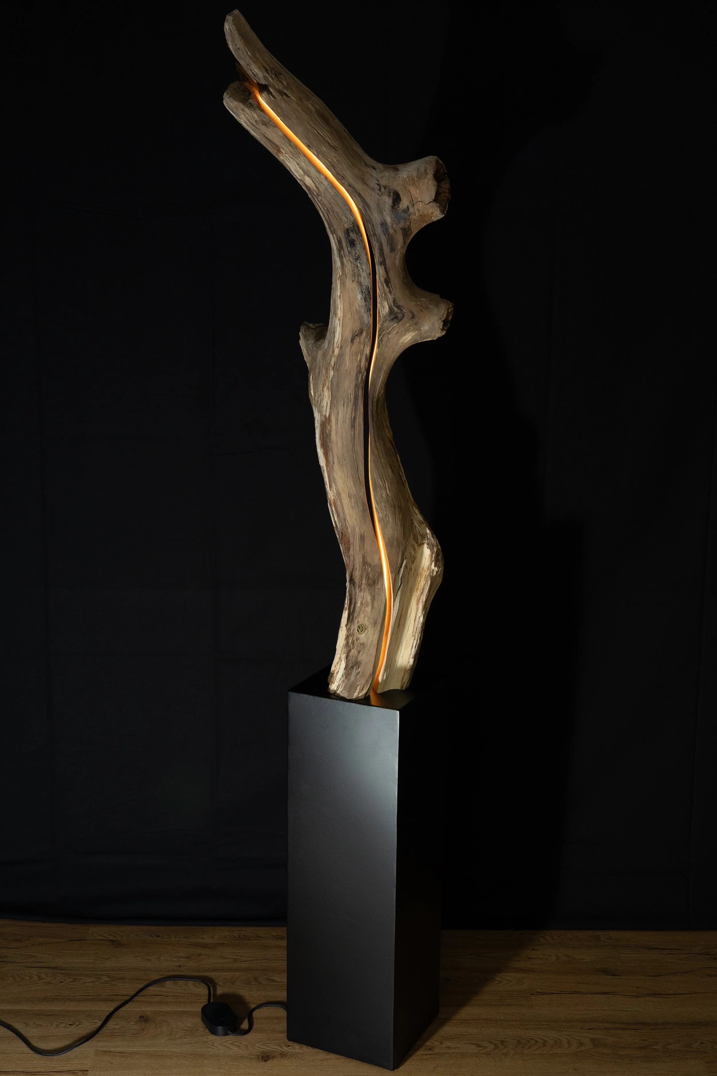 Eine Skulptur aus einem natürlichen Holzstück mit eingeritztem Lichtstreifen, auf einem schwarzen Sockel vor einem schwarzen Hintergrund.