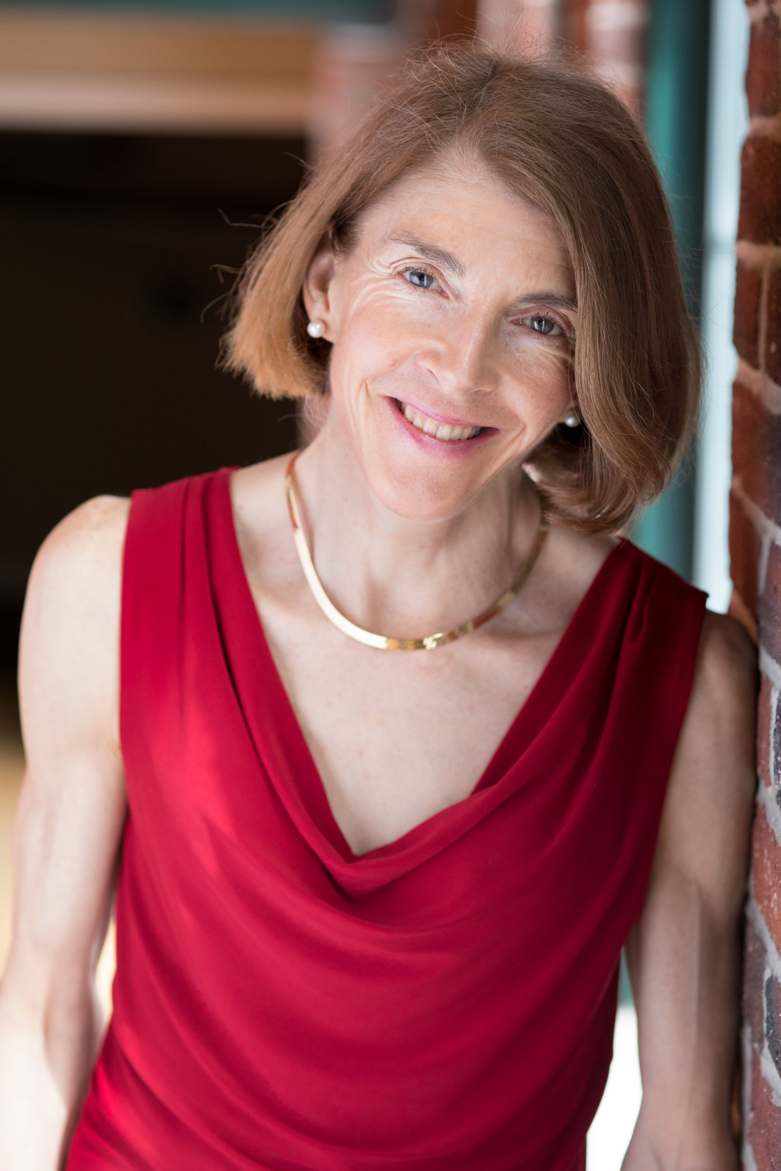 A woman with short red hair and blue eyes, smiling, wearing a red sleeveless dress, pearl earrings, and a gold necklace, leaning against a brick wall.
