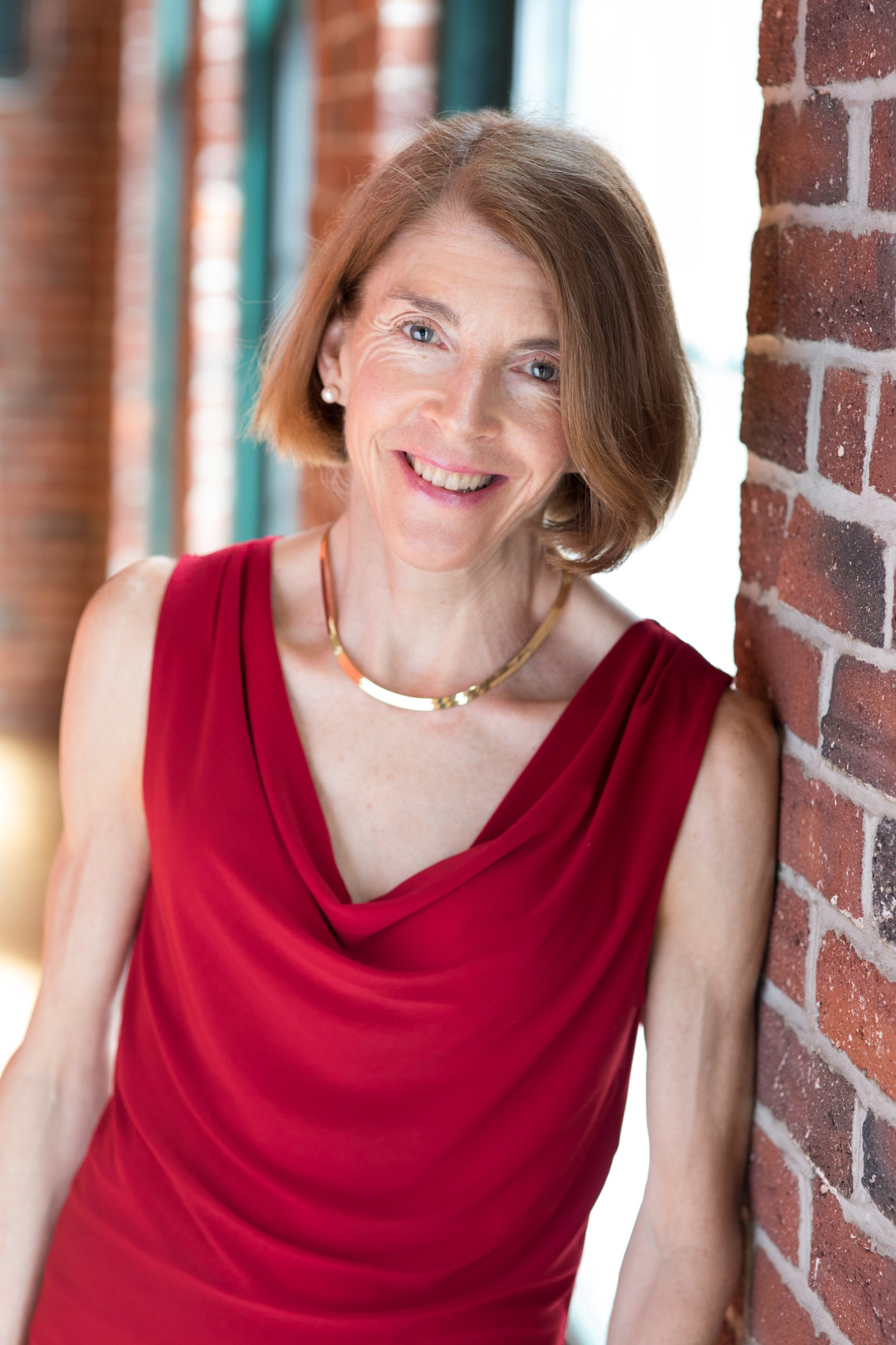 A woman with short red hair smiling, leaning on a brick wall, wearing a sleeveless red dress and a gold necklace.