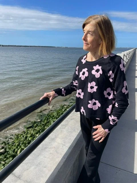 A woman with shoulder-length hair wearing a black sweater with pink and purple floral patterns, standing on a seaside walkway with her hand on a black railing, looking out over the water on a sunny day.