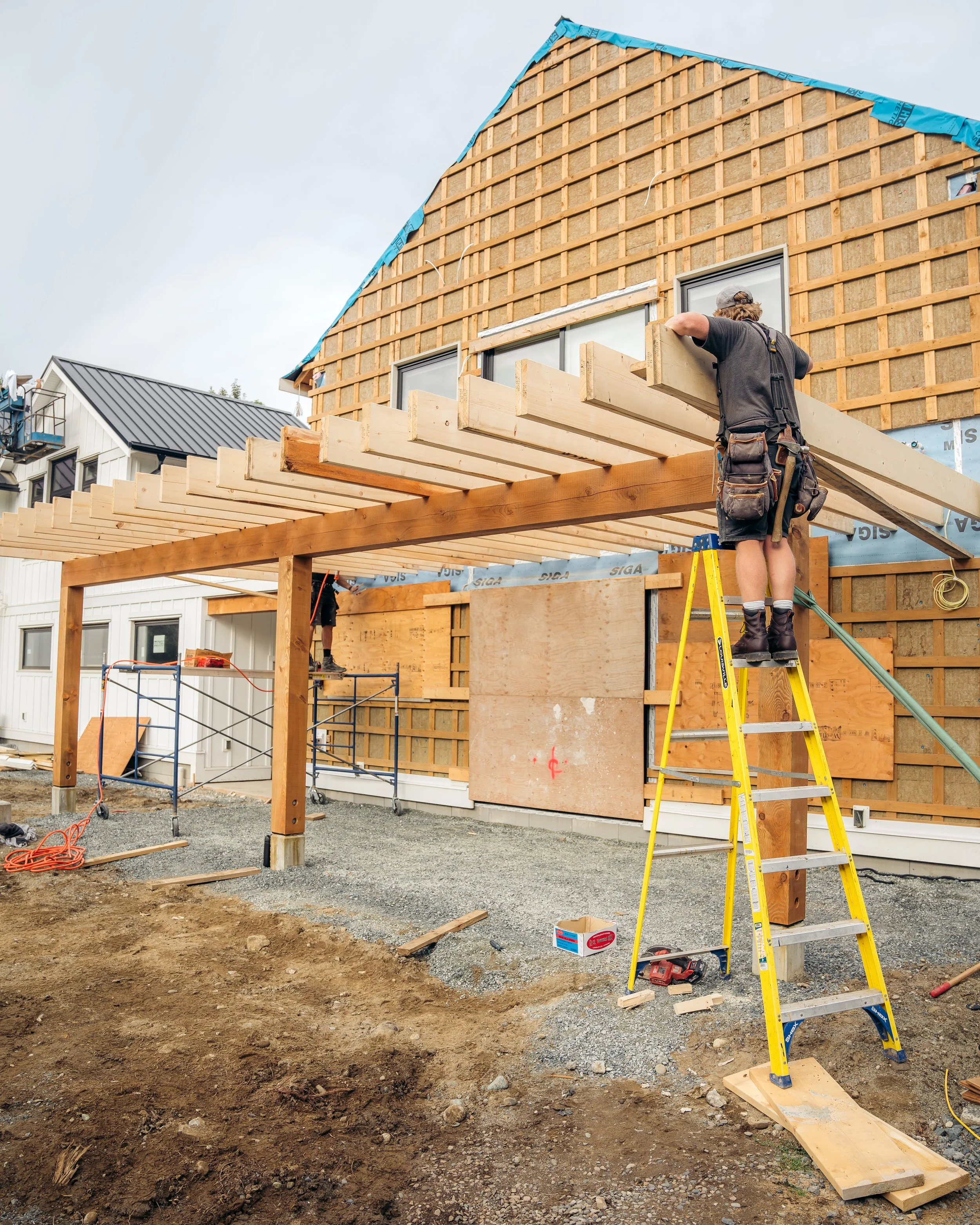 Construction worker on ladder installing a roof on a house under construction, with wooden beams and scaffolding.