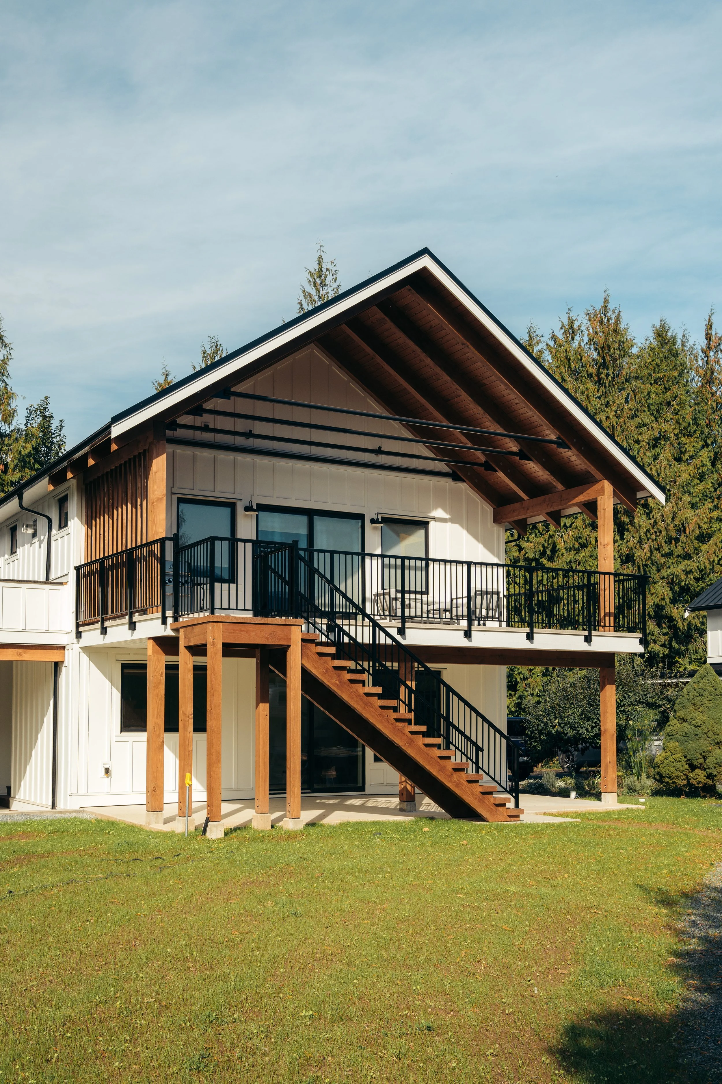Modern two-story house with a spacious balcony and an external staircase, surrounded by a green lawn and trees.
