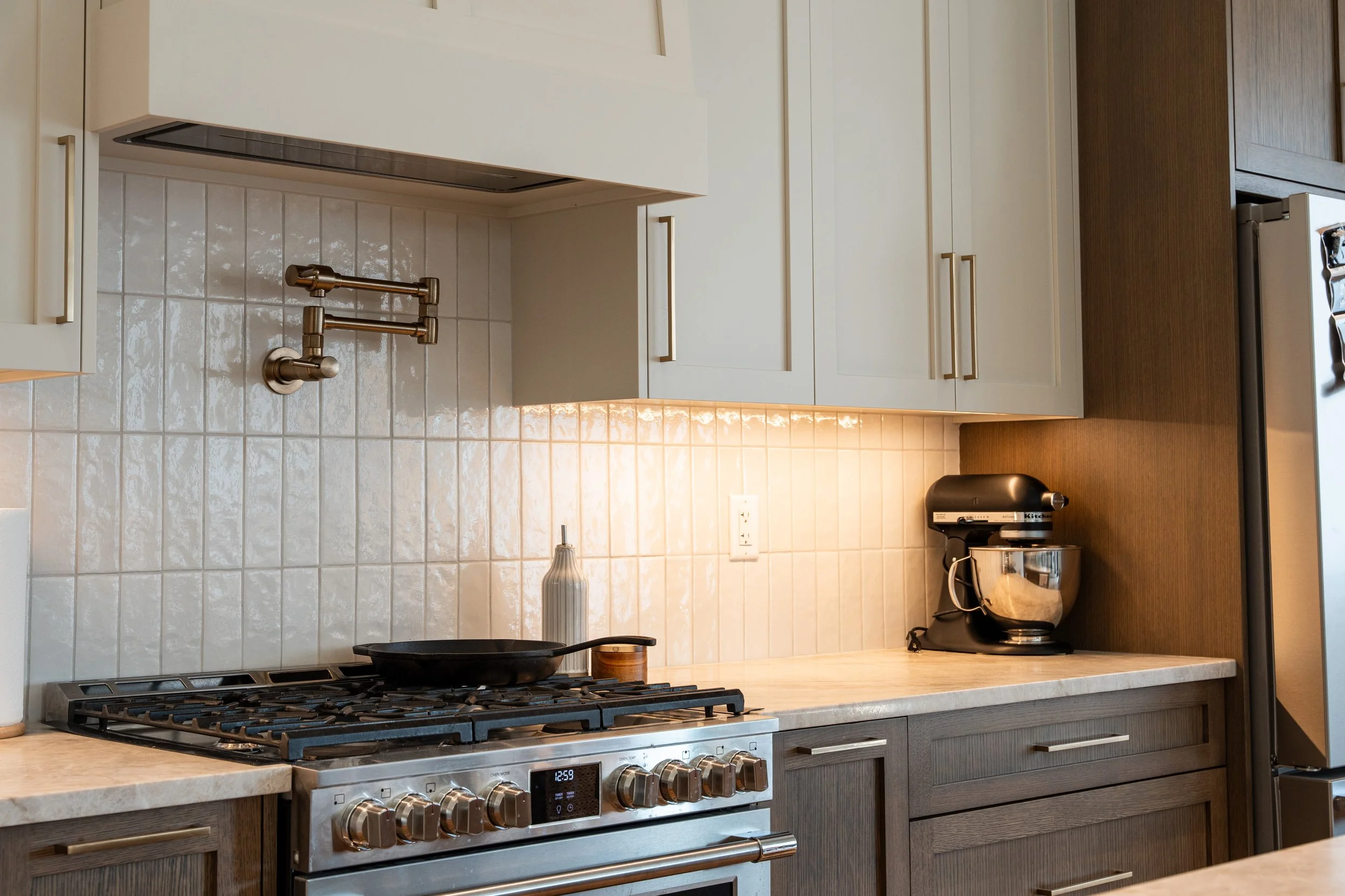 Modern kitchen with white upper cabinets, gray lower cabinets, a stainless steel stove, a black frying pan on the stove, a beige backsplash with tiles, a bottle on the counter, a black KitchenAid mixer, and a silver stand mixer