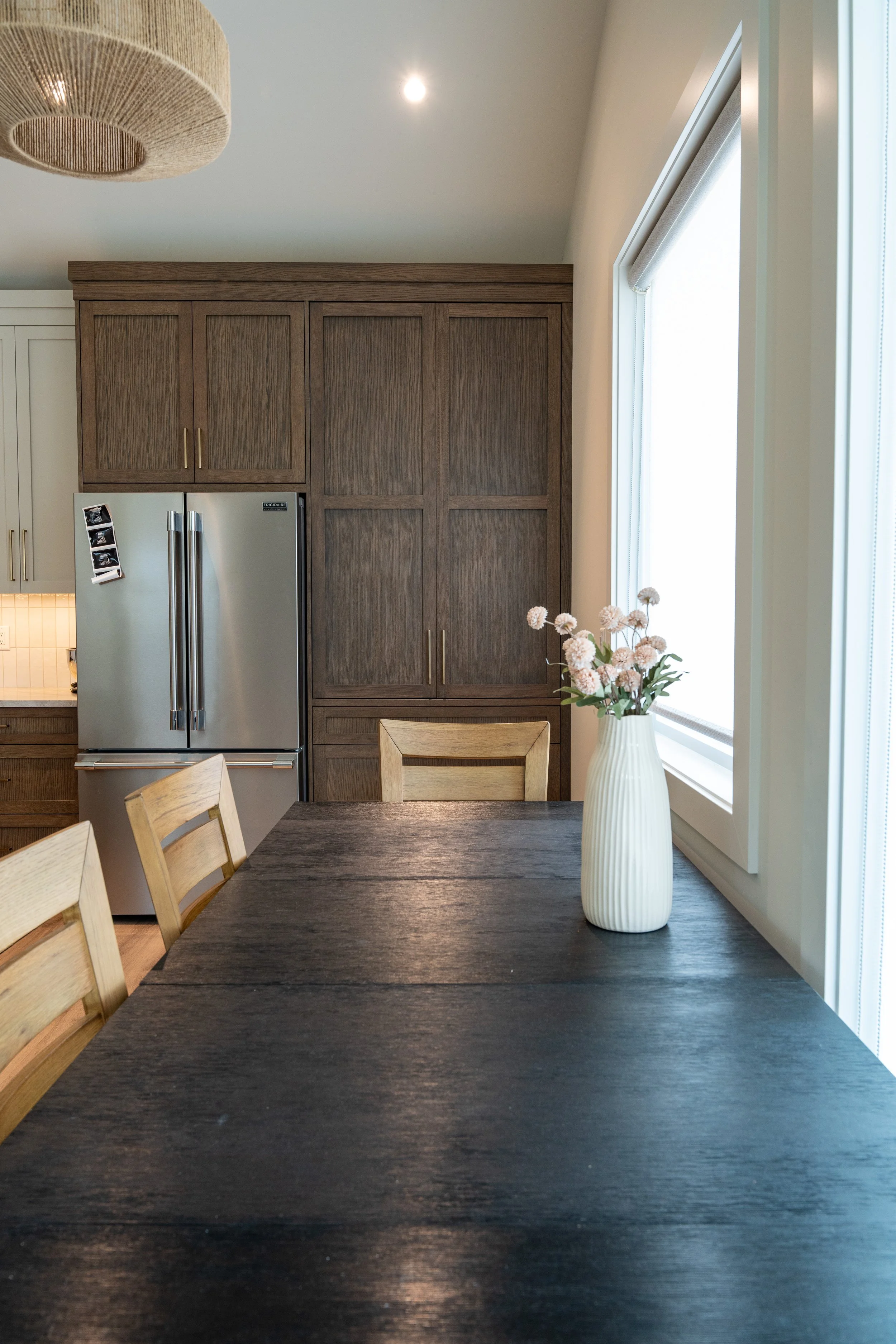 A kitchen dining area with a dark wooden table, beige chairs, a white vase with light pink flowers, and a large window letting in natural light. Wooden cabinets and a refrigerator are visible in the background.